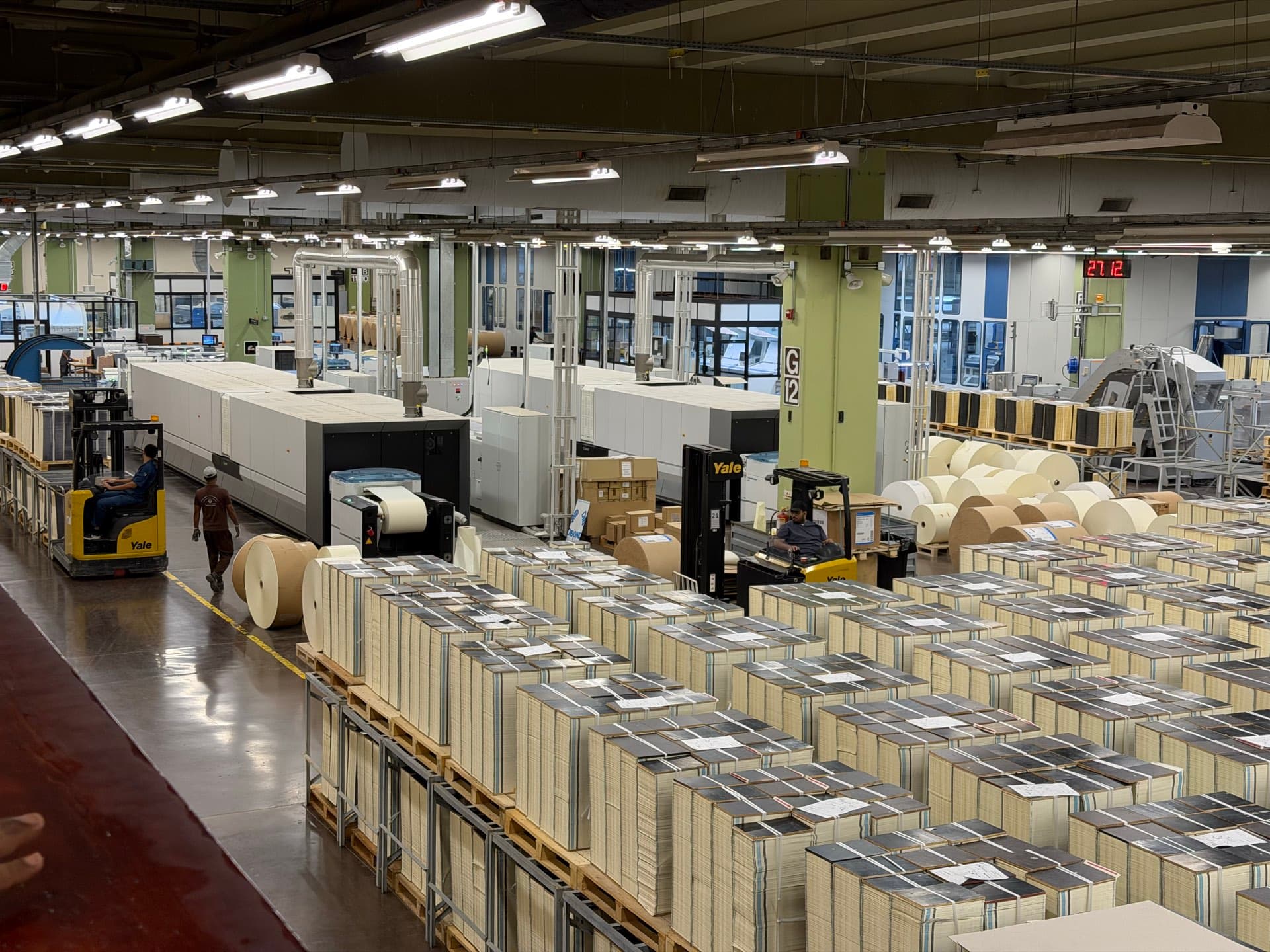 Ranks of pallets stacked with printed Qur’an sheets on the production floor of the King Fahd Complex, with paper reels, a press line and forklift drivers behind them